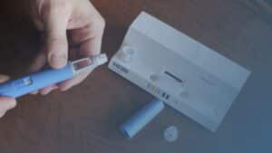 Mãos segurando uma caneta injetora de medicamento azul, com uma agulha visível na ponta, ao lado de uma caixa branca aberta e componentes descartáveis sobre uma mesa de madeira.
