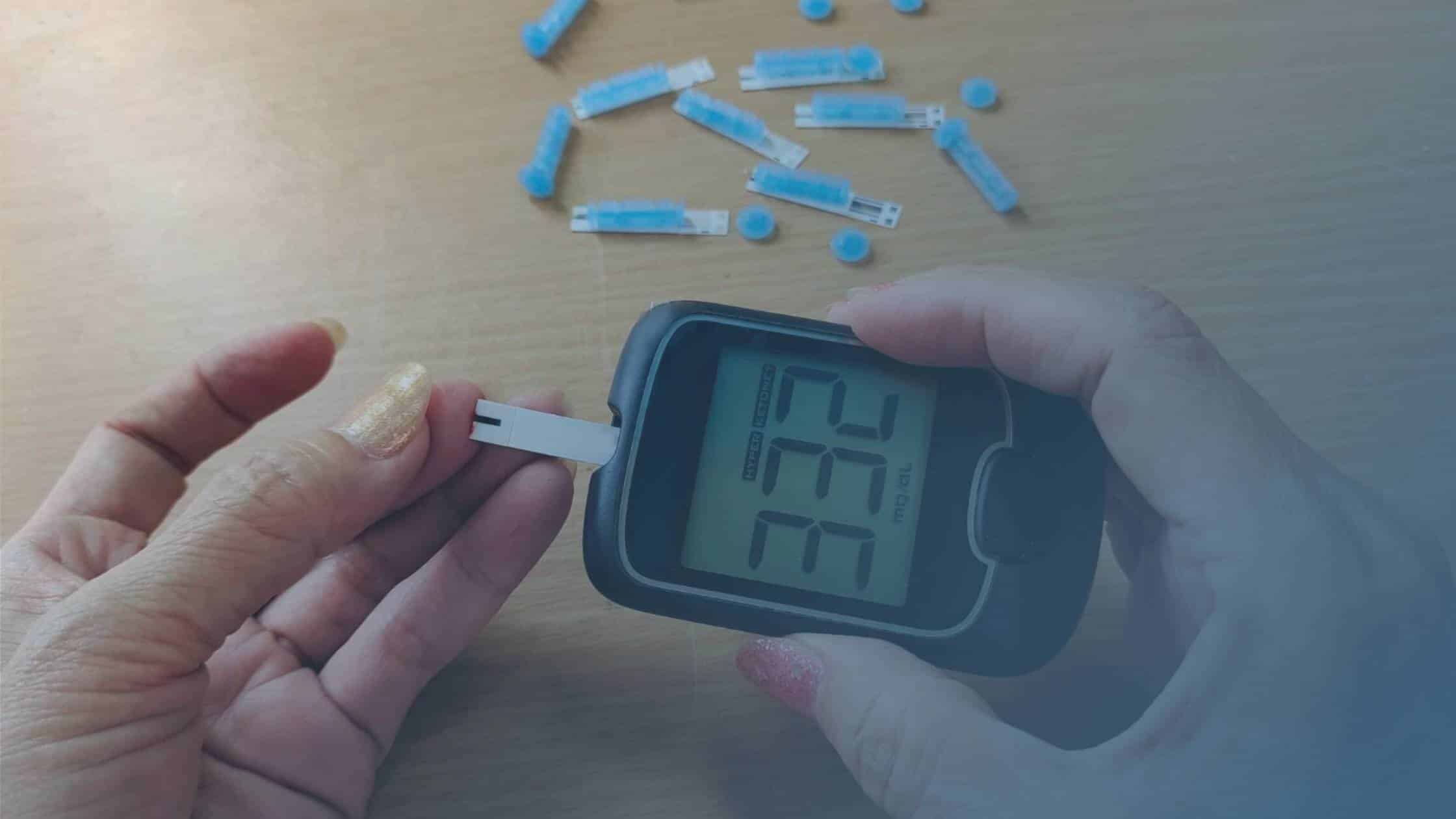Pessoa utilizando um glicosímetro para medir os níveis de glicose no sangue, com tiras de teste ao fundo.